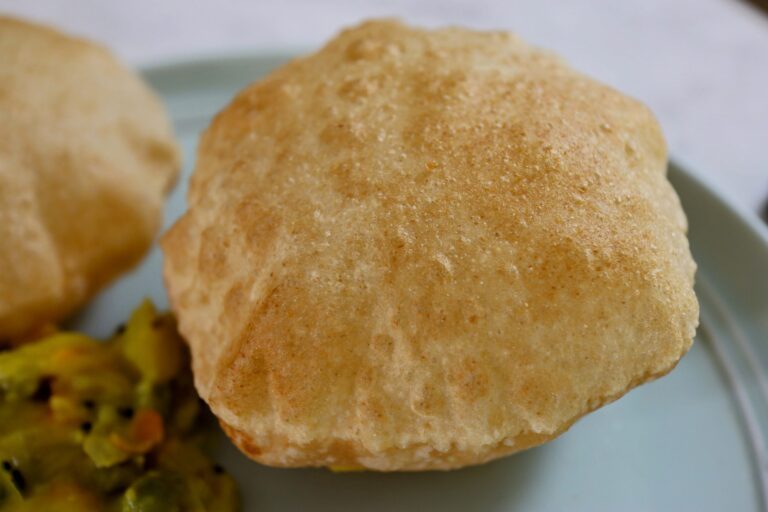 Golden brown, puffy poori bread freshly fried to perfection. One large, round puri is the focus, showing its layered, flaky texture and subtle speckles from frying. Other small pooris are stacked in the background. This popular Indian bread is fried to form air pockets, creating a crisp exterior and soft, pillowy interior.